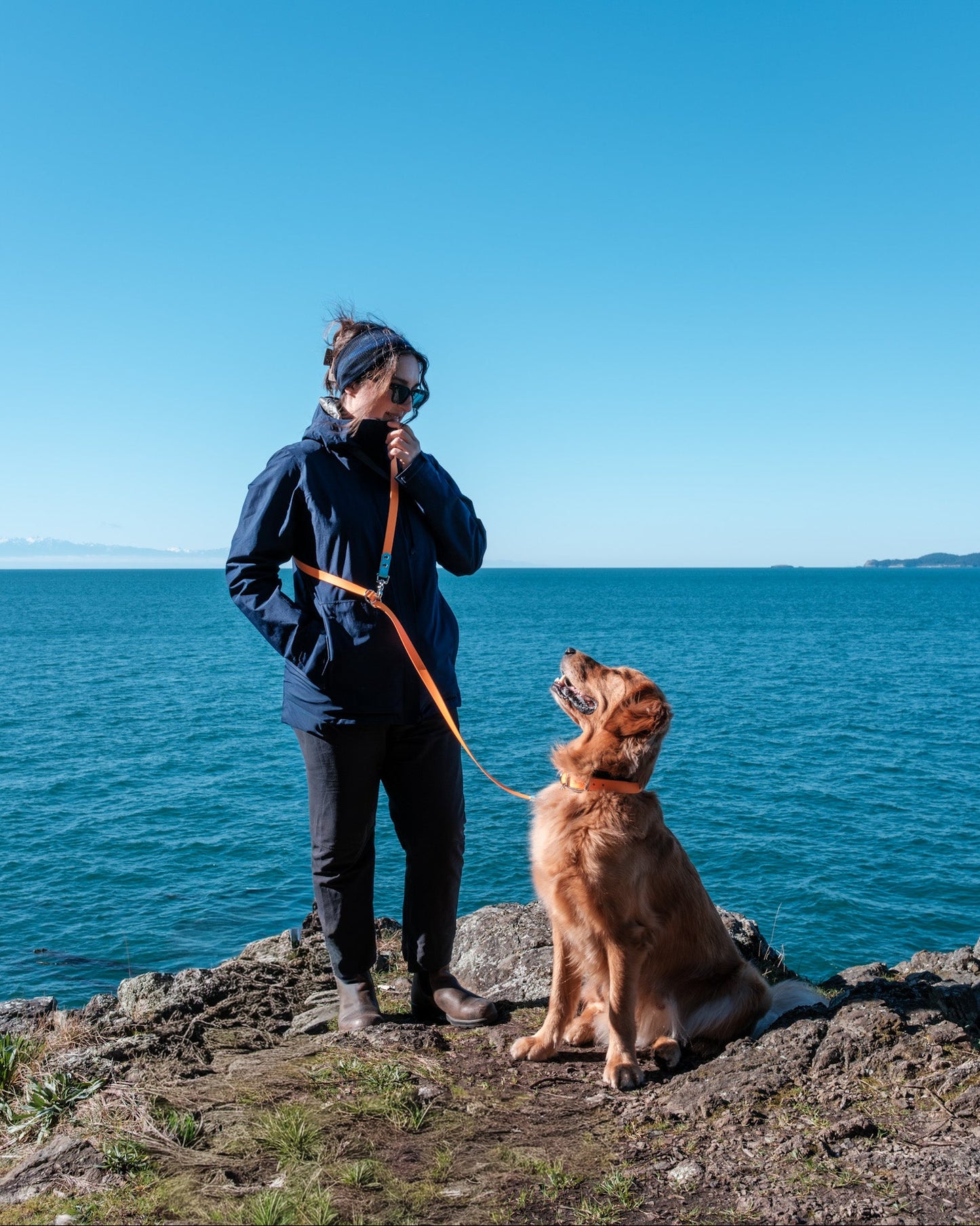 A woman in a navy blue jacket and black pants stands on a rocky shoreline holding an Tangerine Orange and Glacier Blue Multi-Way Leash next to a golden retriever dog, with a bright blue ocean and sky in the background.