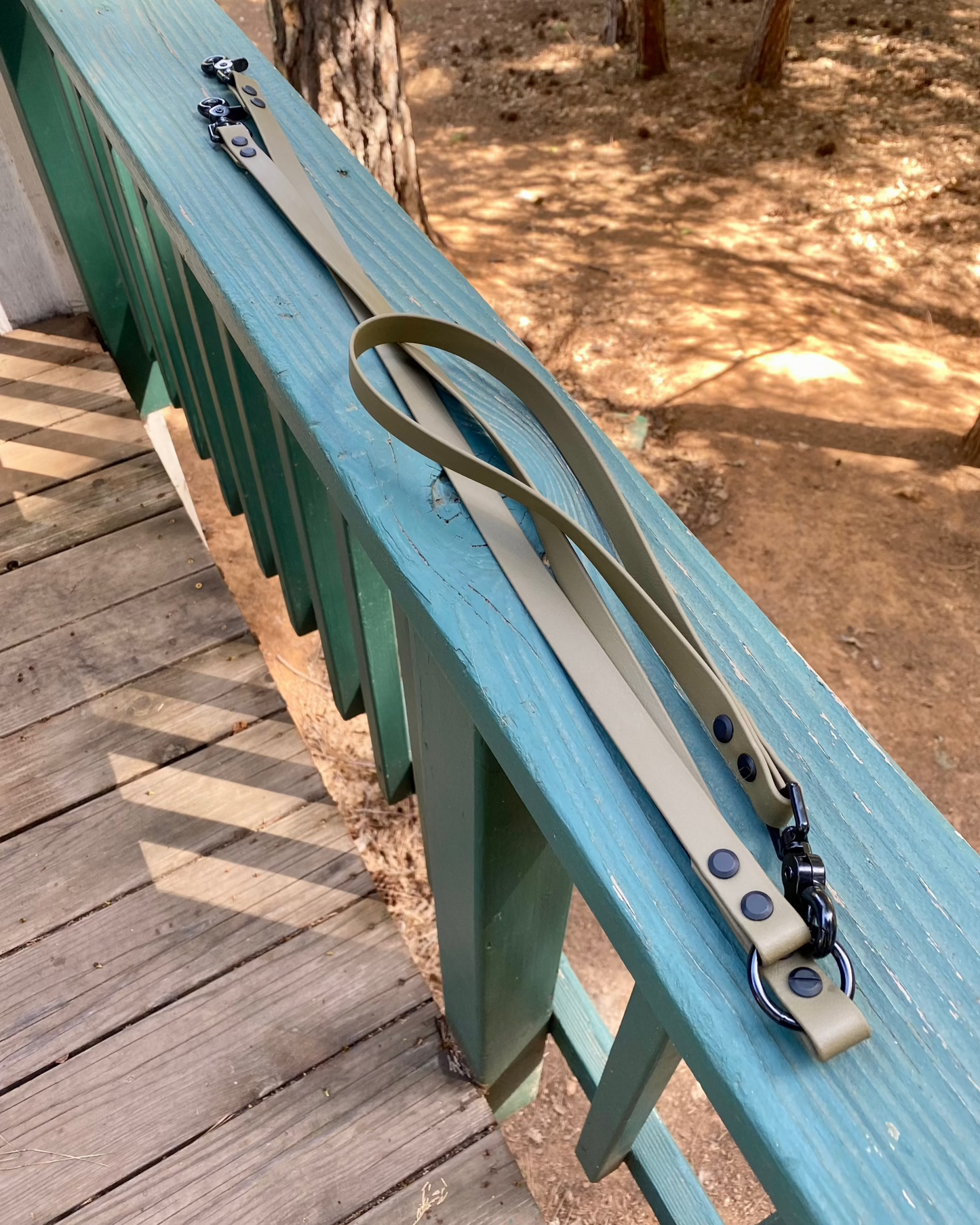 An olive drab two dog leash sitting on a green railing in the woods. 