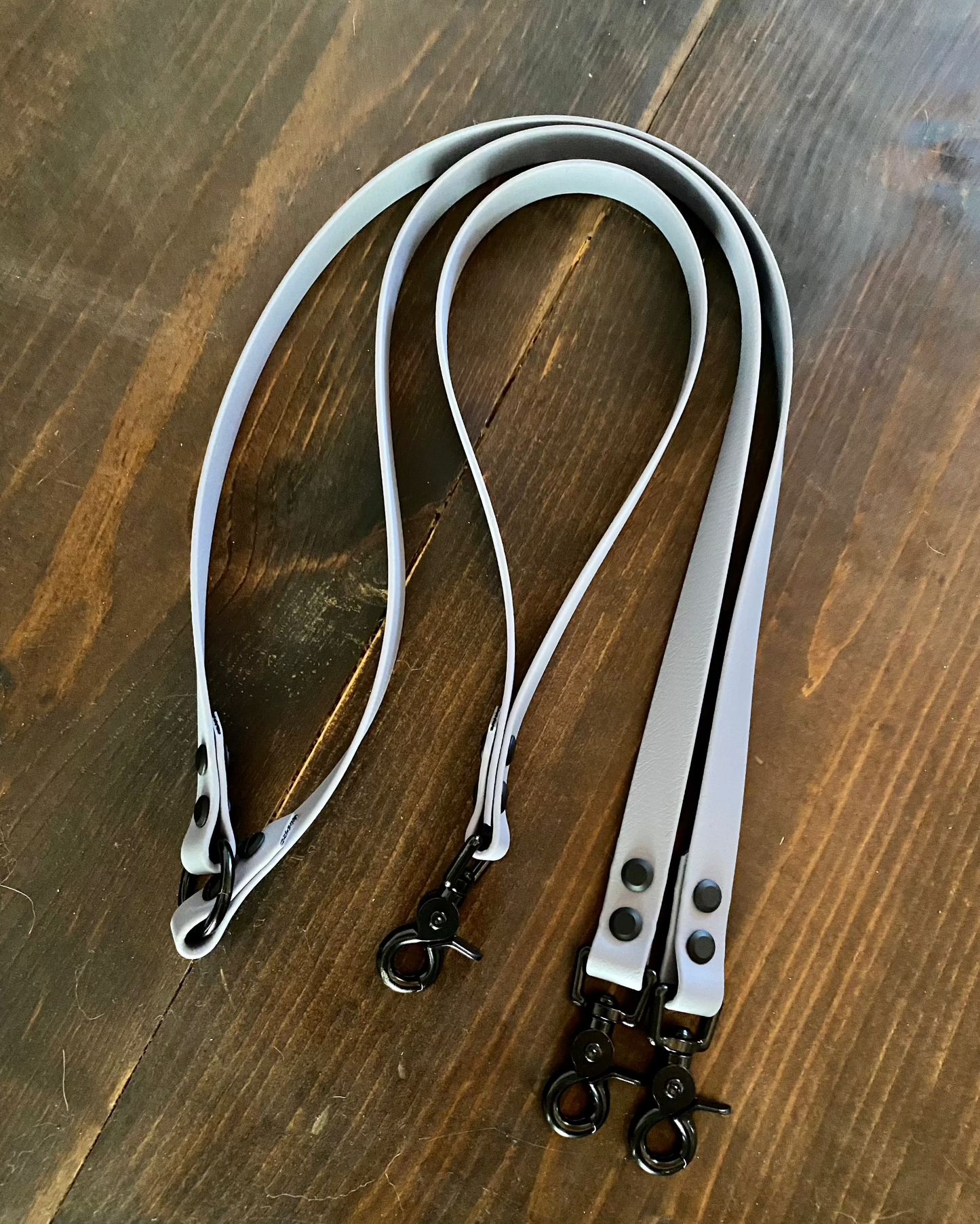 A white two dog leash with a traffic handle attachment laying flat on wood. Both the leash and traffic handle have black hardware.